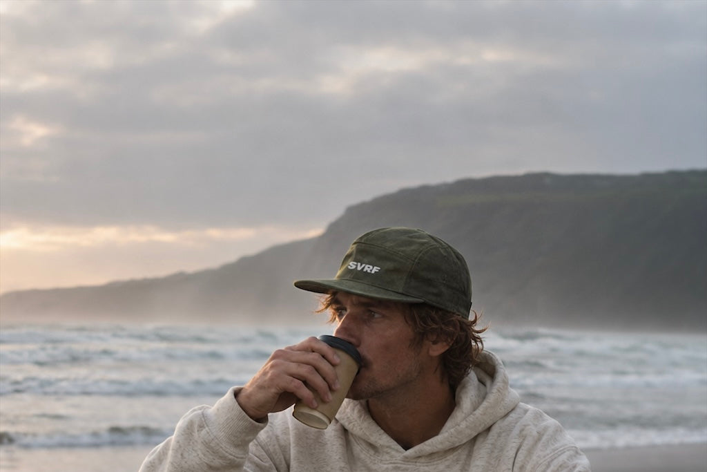 A surfer in an SVRF hoodie and sweatpants sitting on a pebble beach with a flask, grey headland behind
