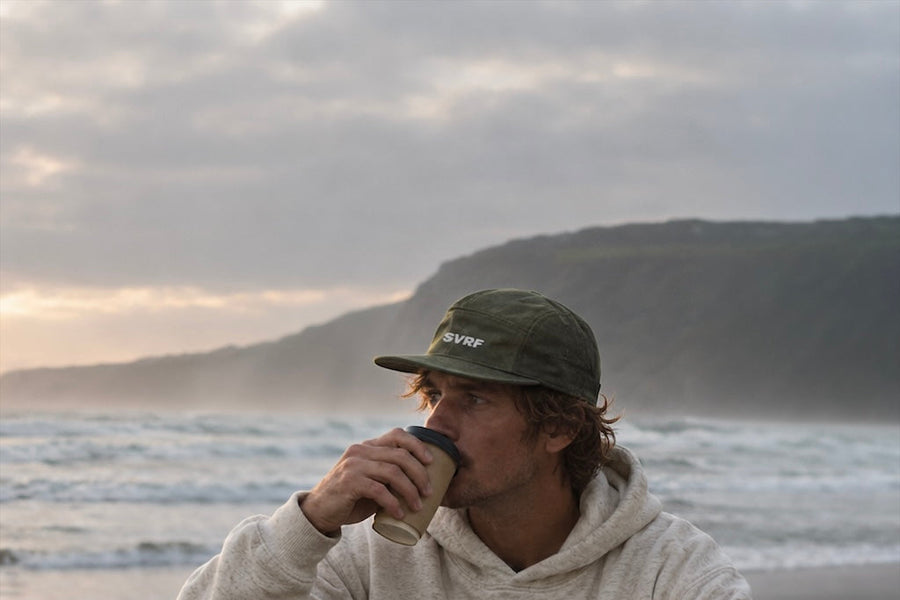 A surfer in an SVRF hoodie and sweatpants sitting on a pebble beach with a flask, grey headland behind