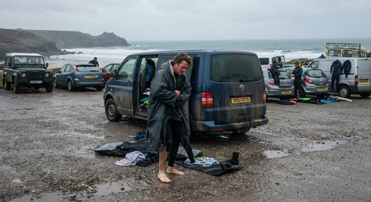 Surfer changing out of a wetsuit beside a van in a coastal car park on a grey UK day