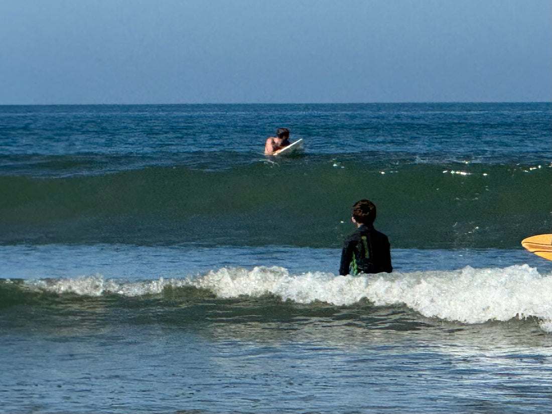 A child in a wetsuit standing in the shorebreak holding a board, another surfer waiting beyond the whitewater