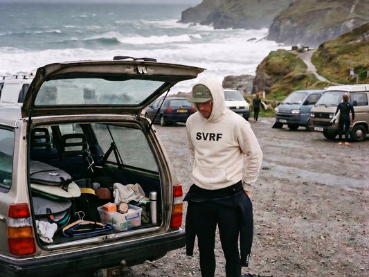 Surfer in an SVRF hoodie peeling a wetsuit down at the open boot of an estate car, grey Cornish surf and cliffs behind