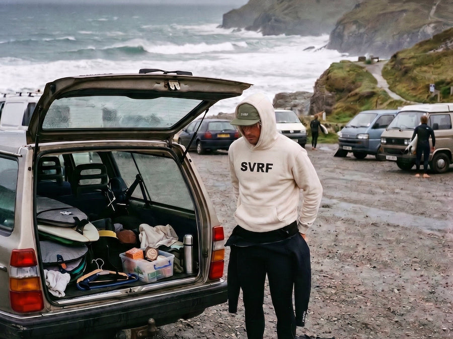 Surfer in an SVRF hoodie peeling a wetsuit down at the open boot of an estate car, grey Cornish surf and cliffs behind