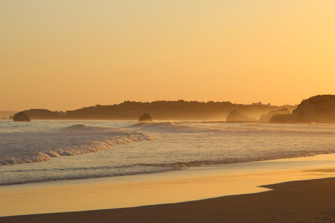 Low golden light over a beach, small waves breaking and rocky headlands in silhouette through the haze