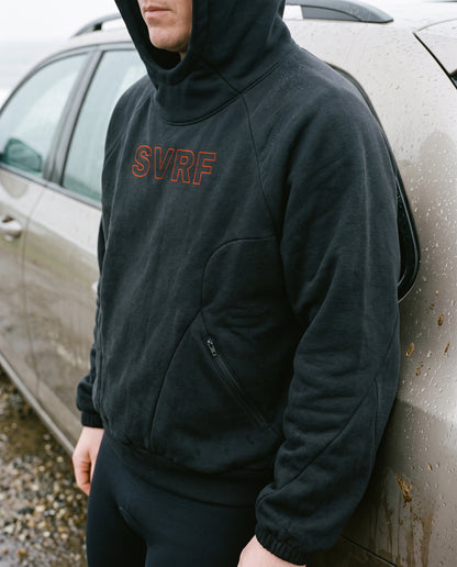 Close crop of the SVRF Raglan Hood in black, hood up, leaning on a muddy car in the rain, showing the raglan seam and zipped front pocket. Render, sample photography to follow