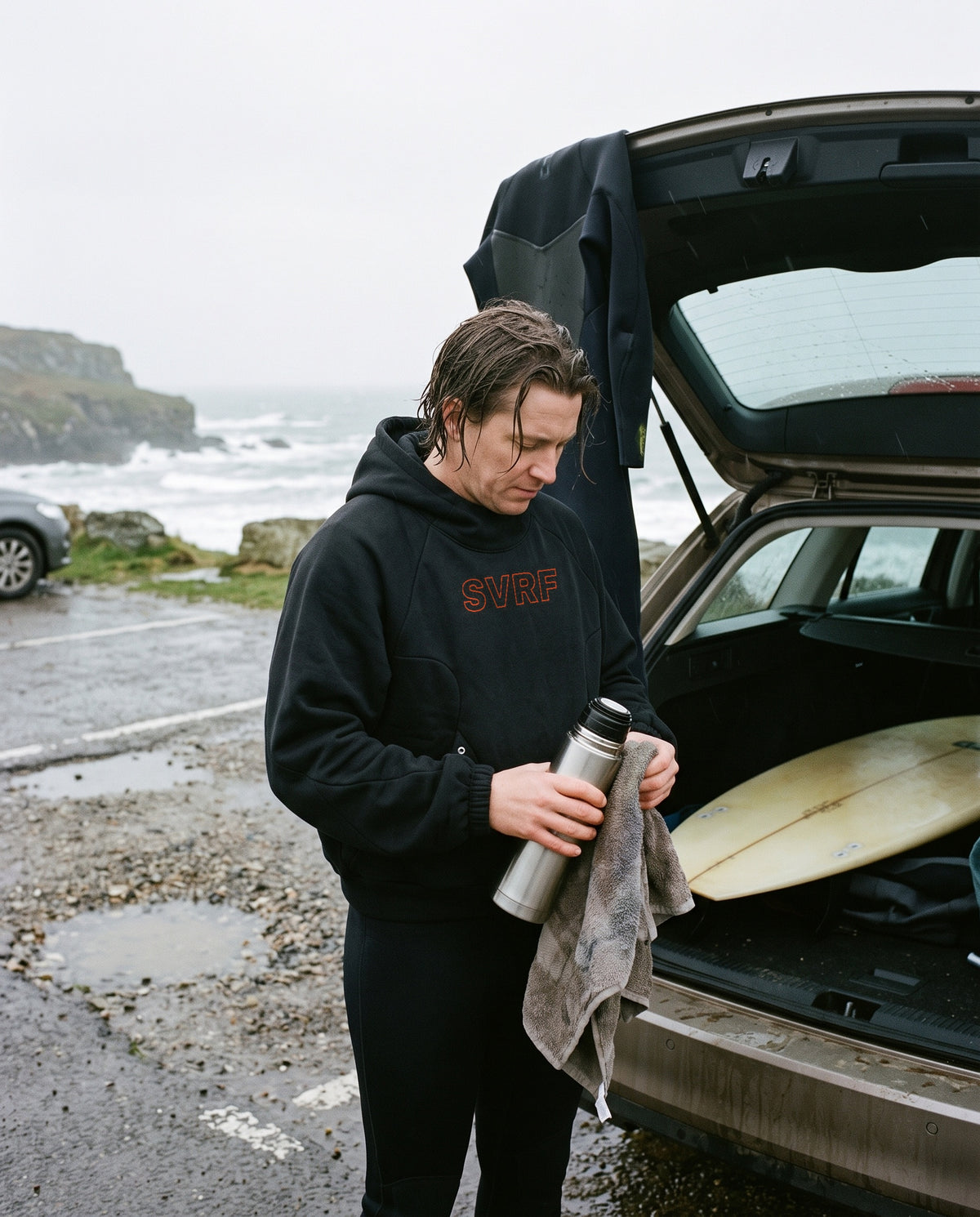 Surfer in the SVRF Raglan Hood in black at an open car boot in a wet Cornish car park, holding a flask and towel, wetsuit hanging off the boot. Render, sample photography to follow