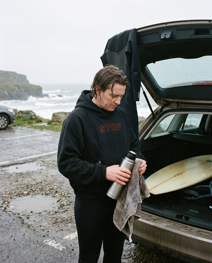 Surfer in the SVRF Raglan Hood in black at an open car boot in a wet Cornish car park, holding a flask and towel, wetsuit hanging off the boot. Render, sample photography to follow